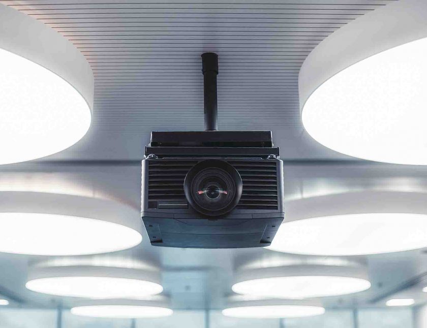 A black modern projector with a plastic case and a huge lens with aberrations in it mounted to a striped ceiling with multiple huge circle ceiling lamps lighting a modern conference hall